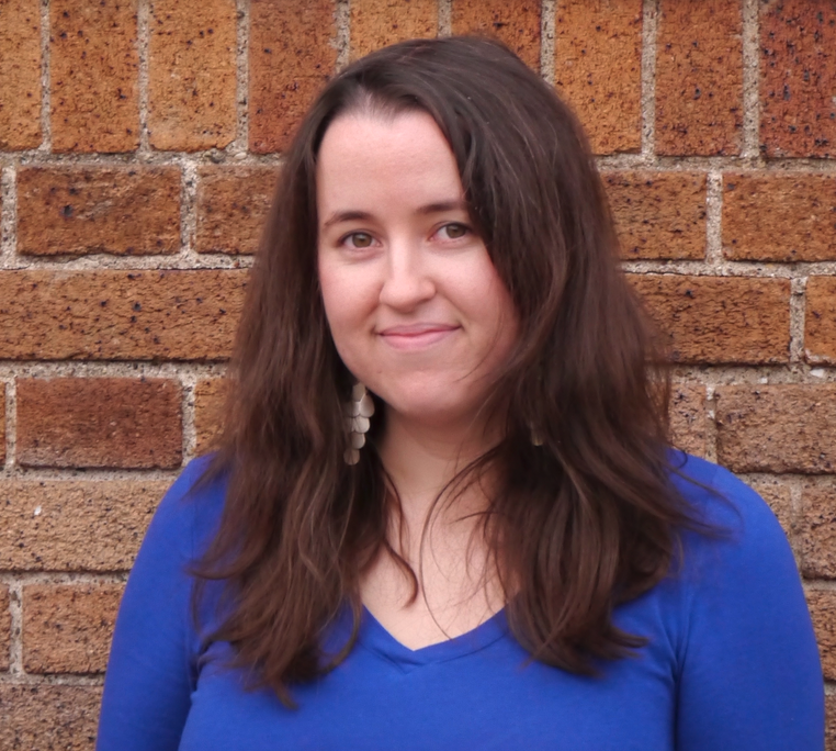 Headshot of Shauna Gordon-McKeon, a white woman with long brown hair in a purple-blue shirt, standing in front of a brick wall